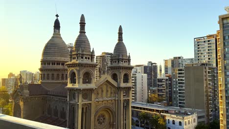 Toma-Panorámica-De-La-Iglesia-Sacramentinos-Rodeada-Por-Los-Rascacielos-De-Santiago-Al-Atardecer,-Chile