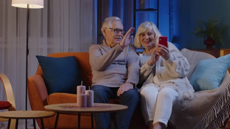 Smiling-senior-couple-grandmother-grandfather-making-video-call-online-on-mobile-phone-at-home-sofa