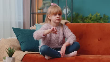 Child-girl-kid-sitting-on-sofa-at-home-alone-showing-thumbs-down-sign-gesture,-dislike,-disapproval