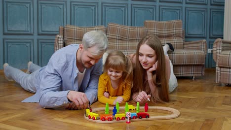 Family-of-mother,-father-with-daughter-child-girl-riding-toy-train-on-wooden-railway-at-home-room