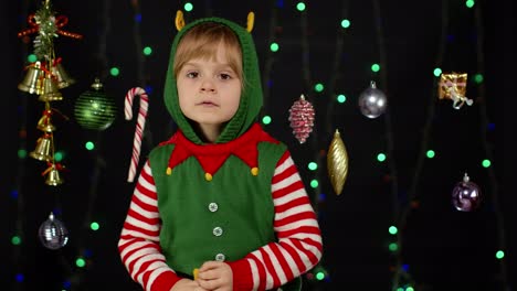 Kid-girl-in-Christmas-elf-Santa-Claus-helper-costume-looks-at-camera-makes-big-eyes-covering-mouth