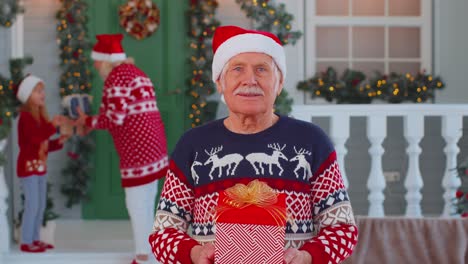 Portrait-of-grandfather-man-presenting-gift-box-smiling-near-decorated-Christmas-house-with-family
