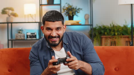 Indian-man-using-joystick-controller-playing-video-console-winning-online-fun-game-sits-on-home-sofa