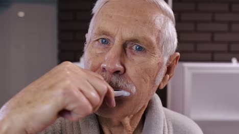 Senior-man-grandfather-in-bathrobe-brushing-teeth,-looking-into-mirror.-Morning-hygiene-at-bathroom