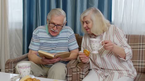 Lächelndes-älteres-Paar,-Großvater-Und-Großmutter-Ruhen-Sich-Auf-Dem-Sofa-Aus,-Trinken-Kaffee-Und-Lesen-Zu-Hause-Ein-Buch