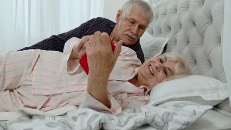 Senior-elderly-couple-wearing-pajamas-lying-on-bed-looking-on-mobile-phone-laughing-and-having-fun