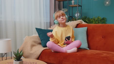 Child-girl-kid-with-smartphone-in-headphones-dancing-singing-listening-music-at-home-alone-on-sofa