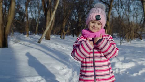 Joyful-little-child-girl-smiling,-showing-thumbs-up-gesture-on-snowy-road-in-winter-park-outdoors
