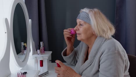 Old-senior-woman-grandmother-taking-care-of-skin,-applying-anti-wrinkle-foundation-with-sponge