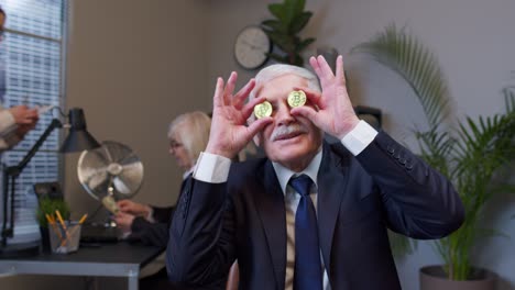 Elderly-man-boss-dancing,-fooling-around,-making-silly-faces-with-bitcoin-coints-eyes-in-office