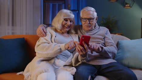 Una-Feliz-Pareja-De-Ancianos-Sonriente-Con-Un-Smartphone-En-El-Sofá-De-Casa-Pasa-Tiempo-Libre-En-Las-Redes-Sociales