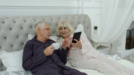 Senior-elderly-couple-wearing-pyjamas-lying-on-bed-looking-on-digital-tablet-making-online-shopping