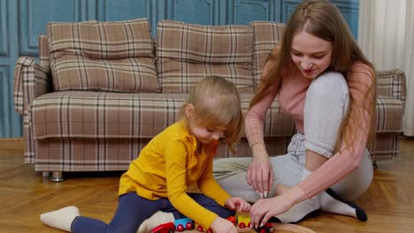 Mother-with-little-daughter-child-girl-riding-toy-train-on-wooden-railway-blocks-board-game-at-home