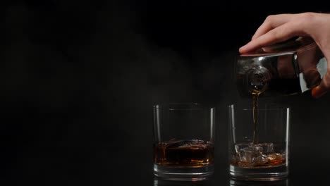 Barman-pouring-whiskey,-cognac,-brandy-from-bottle-into-glasses-with-ice-cubes-on-black-background