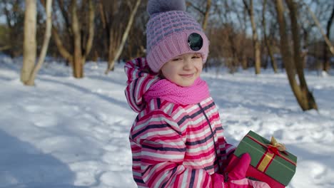 Smiling-child-kid-in-snowy-winter-forest-park-looking-at-camera,-holding-Christmas-present-gift-box