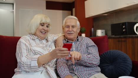 Glückliches-Lächelndes-älteres-Paar-Mit-Mobiltelefon-Zu-Hause.-Ausruhen-Auf-Dem-Sofa-Im-Gemütlichen-Wohnzimmer