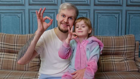 Father-and-little-child-daughter-kid-in-pajamas-sit-on-couch-in-room-smiling-showing-ok-gesture-sign