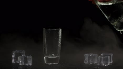 Barman-hand-pours-vodka,-tequila-or-sake-from-bottle-into-glass-on-black-background-with-ice-cubes