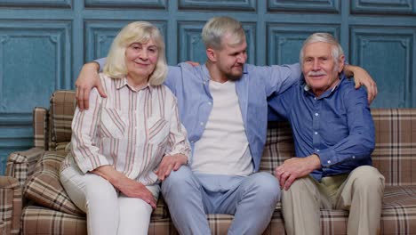 Family-of-senior-grandparents-with-adult-son-embracing-having-fun-looking-at-camera-bonding-at-home
