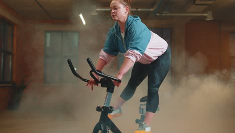 Athletic-girl-performing-aerobic-riding-training-exercises-on-cycling-stationary-bike-in-foggy-gym