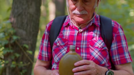 Tired-thirsty-senior-old-Caucasian-hiker-grandfather-relaxing,-drinking-water-in-forest-after-walk