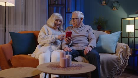 Senior-couple-grandfather-grandmother-making-online-shopping-on-mobile-phone-sitting-at-home-sofa