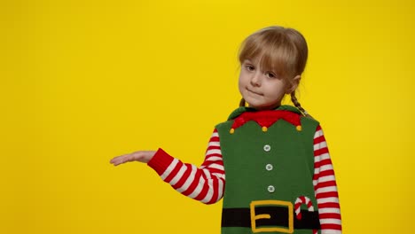 Kid-girl-in-Christmas-elf-Santa-helper-costume-points-fingers-at-blank-space-shows-advertising-area