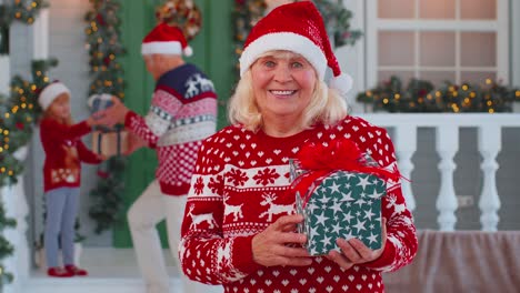 Portrait-of-grandmother-woman-presenting-gift-box-smiling-near-decorated-Christmas-house-with-family