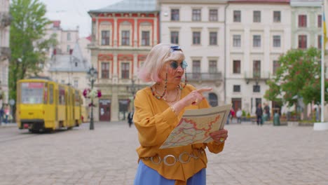 Senior-stylish-tourist-granny-woman-walking-along-street,-looking-for-way-using-paper-map-in-city
