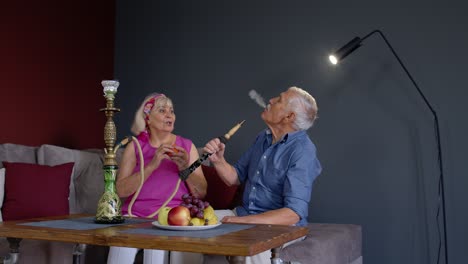 Senior-couple-smoking-hookah-eating-fruits-celebrating-retirement-anniversary-in-living-room-at-home