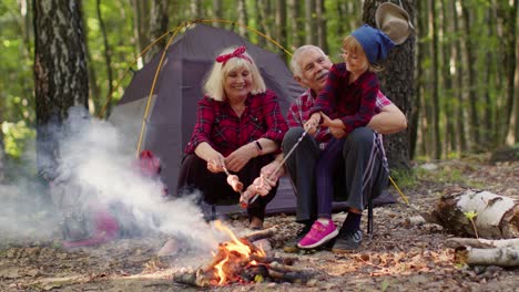 Senior-grandmother-grandfather-with-granddaughter-cooking-frying-sausages-over-campfire-in-wood