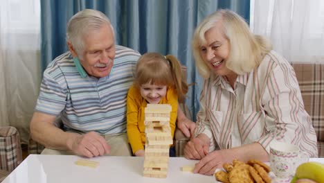 Senior-couple-grandparents-and-granddaughter-enjoying-board-game-building-tower-from-blocks-at-home