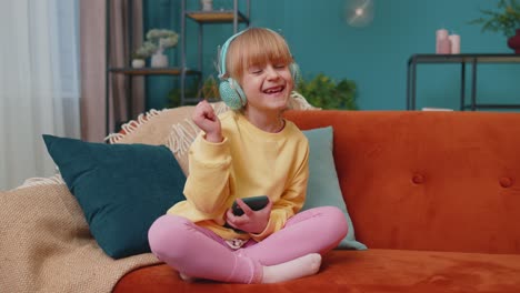 Child-girl-kid-with-smartphone-in-headphones-dancing-while-listening-music-at-home-alone-on-sofa
