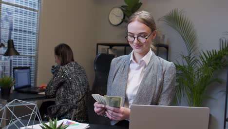 Cheerful-rich-business-woman-manager-counting-money-dollar-cash-after-working-on-laptop-at-office