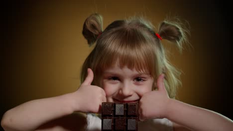 Little-child-girl-kid-eating-chocolate-bar-dessert,-showing-thumbs-up-isolated-on-dark-background