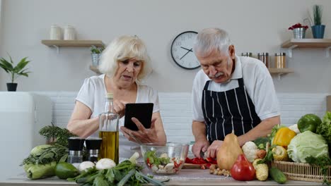 Vegan-senior-couple-cooking-salad-with-raw-vegetables.-Looking-on-digital-tablet-for-online-recipe