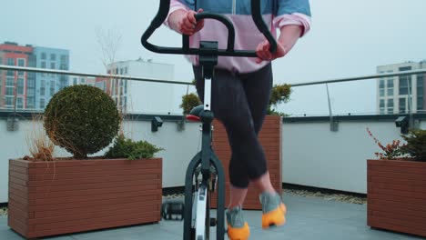 Sportliches-Mädchen,-Das-Aerobic-Trainingsübungen-Auf-Einem-Stationären-Fahrrad-Auf-Dem-Hausdach-Durchführt