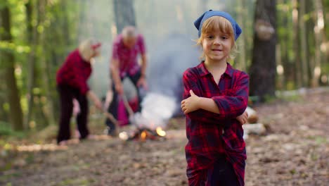 Child-girl-kid-traveler-tourist-on-camping-looking-at-camera-showing-thumbs-up-near-bonfire-in-wood