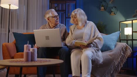 Senior-grandparents-couple-reading-book,-using-laptop-pc-on-couch-in-night-living-room-at-home