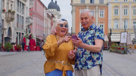 Senior-old-stylish-tourists-looking-smartphone-sincerely-rejoicing-win,-message-with-good-news