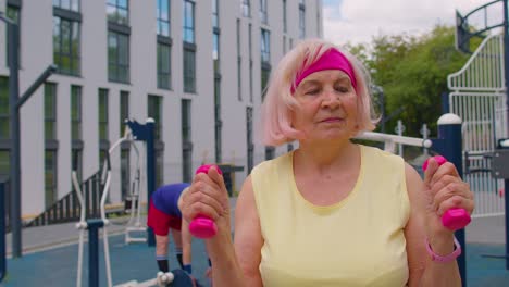 Senior-woman-grandmother-athlete-doing-routine-weightlifting-dumbbells-exercises-on-playground