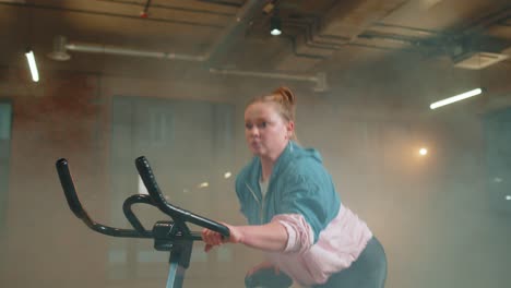 Athletic-woman-riding-on-spinning-stationary-bike-training-routine-in-haze-gym,-weight-loss-indoors