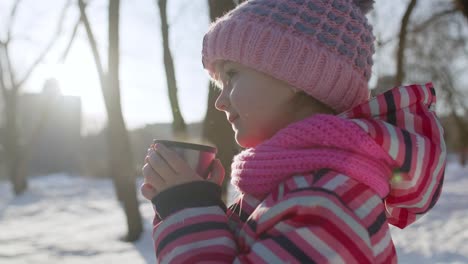 Smiling-child-kid-girl-drinking-hot-drink-tea-from-cup,-trying-to-keep-warm-in-winter-park-forest