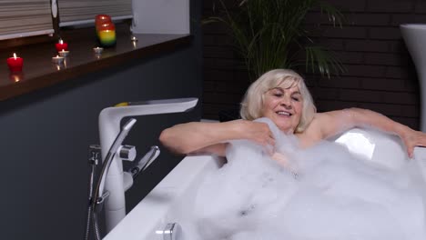 Sexy-senior-elderly-blonde-woman-grandmother-is-taking-foamy-bath-in-luxury-bathroom-with-candles