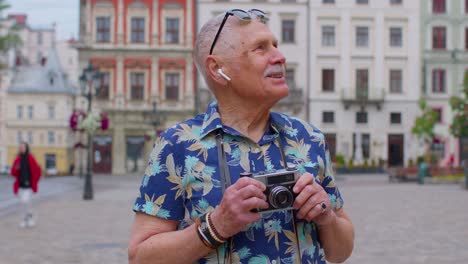 Senior-man-tourist-with-retro-photo-camera,-smiling,-listening-music-earphones,-dancing-on-street