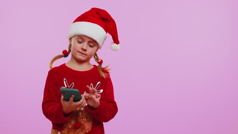 Girl-toddler-in-Christmas-Santa-hat-with-mobile-phone-showing-pointing-empty-place,-advertising-area