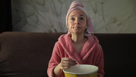 Woman-watching-a-late-night-movie-at-TV,-eating-popcorn.-Bathrobe,-facial-mask