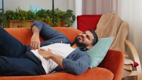 Indian-man-lying-on-sofa-feeling-sudden-strong-abdominal-stomach-ache-gastritis-poisoning-diarrhea