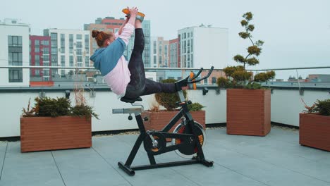 Athletic-woman-makes-twine-training-routine-on-spinning-stationary-bike-on-house-roof,-weight-loss