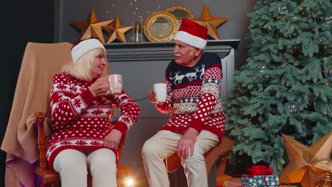 Ältere-Frau-Und-Ehemann-Unterhalten-Sich-Und-Trinken-Tee-Neben-Einem-Leuchtenden-Weihnachtsbaum-Zu-Weihnachten-Im-Wohnzimmer